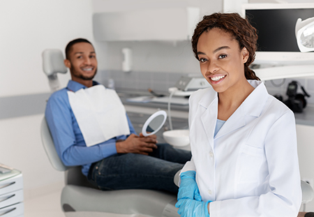 A dental hygienist wearing a white coat and holding a dental mirror, with a smiling man sitting in a dental chair nearby.