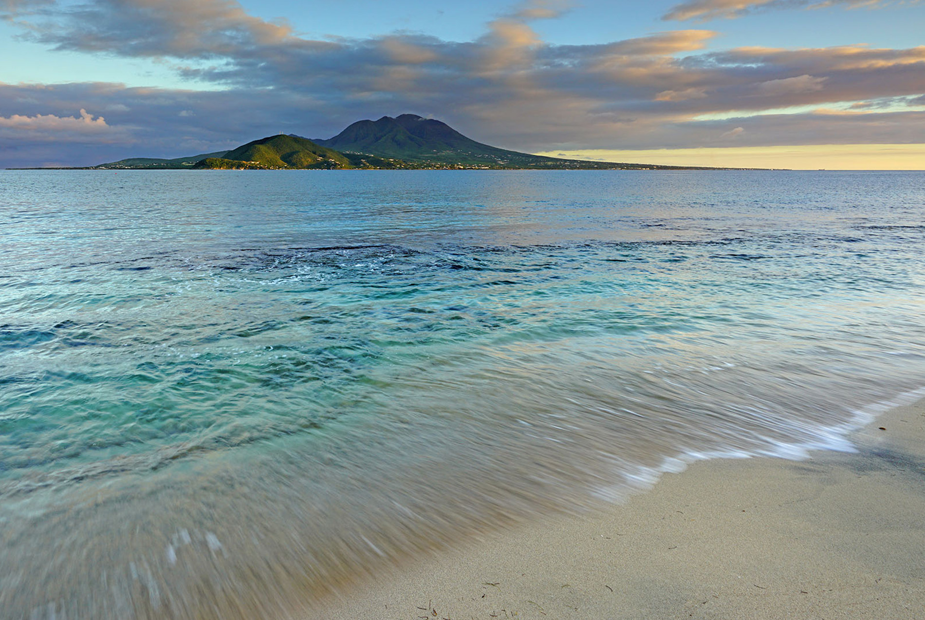 A serene beach scene with clear blue waters, white foam on the waves, and a tropical island with lush vegetation in the distance under a partly cloudy sky.