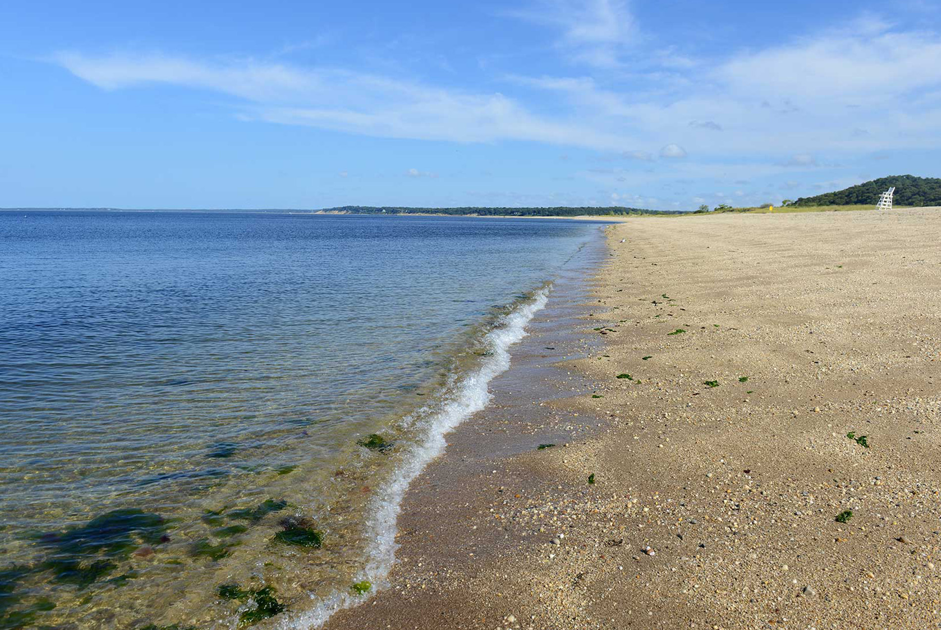 The image shows a sandy beach with water at low tide, adjacent to a body of water, possibly an ocean, under a clear blue sky with some scattered clouds.