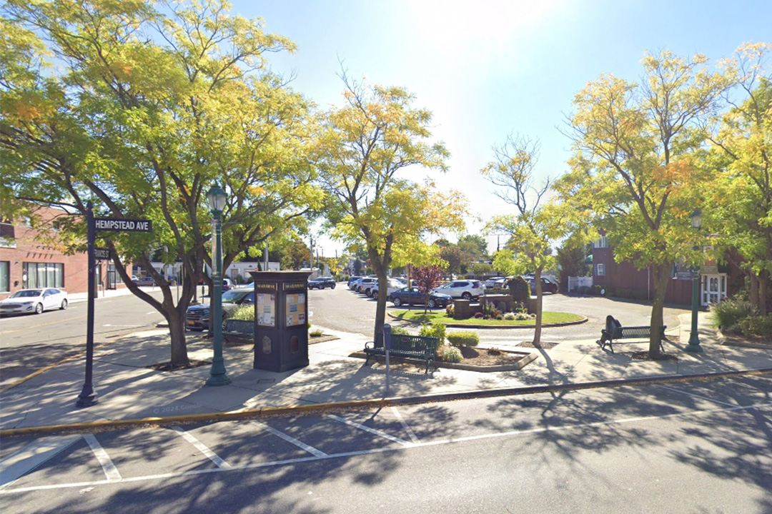 The image shows a sunny day with a street corner featuring trees, parked cars, benches, and a clear blue sky.