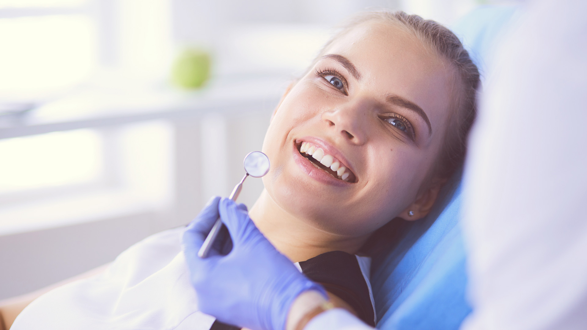 The image shows a woman sitting in a dental chair with a smiling expression, receiving dental care from a professional wearing protective gloves, while the background suggests a clean, modern dental office setting.