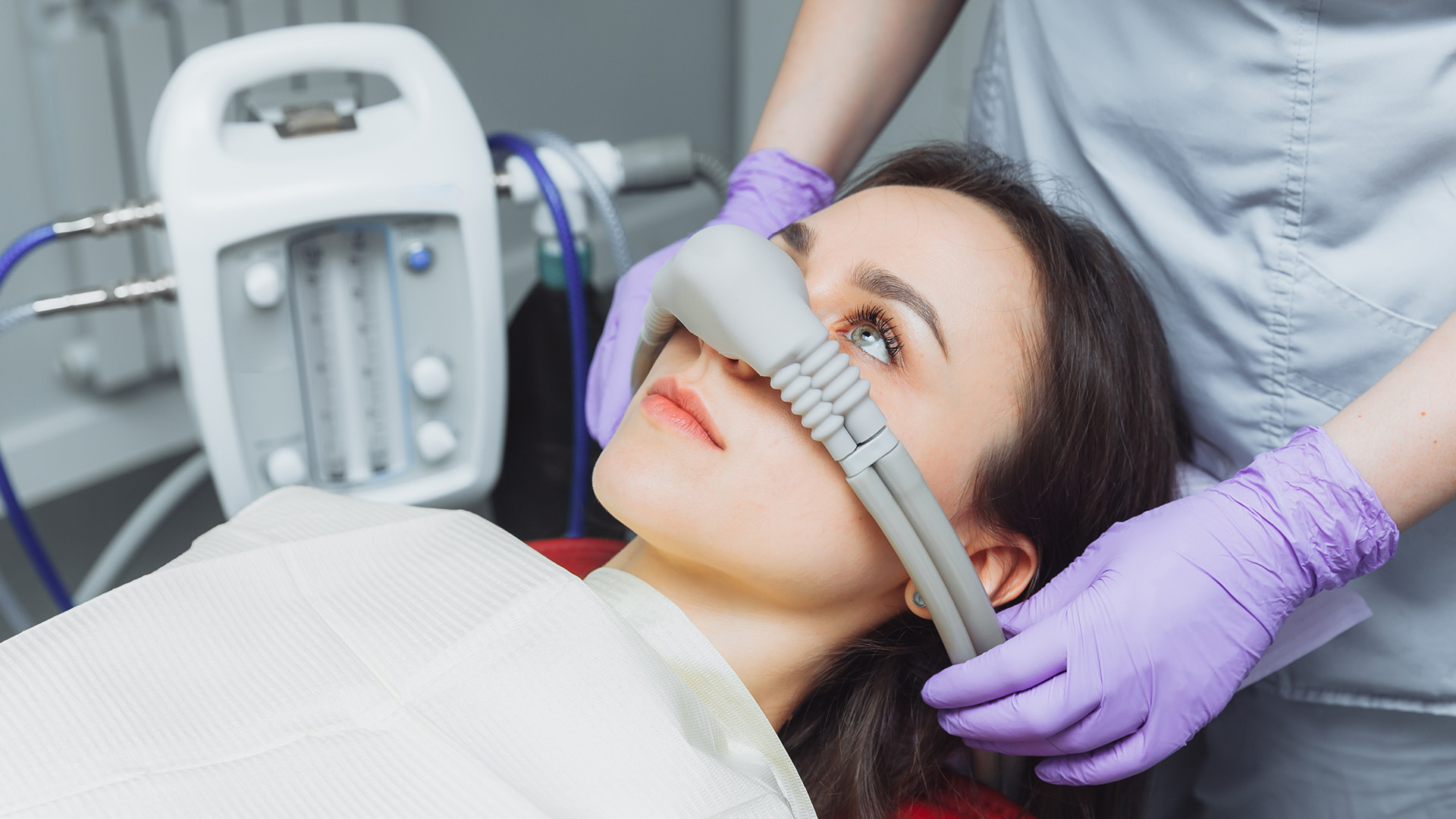 A woman receiving dental treatment with a device attached to her face while wearing a surgical gown and a medical professional using a computer monitor nearby.