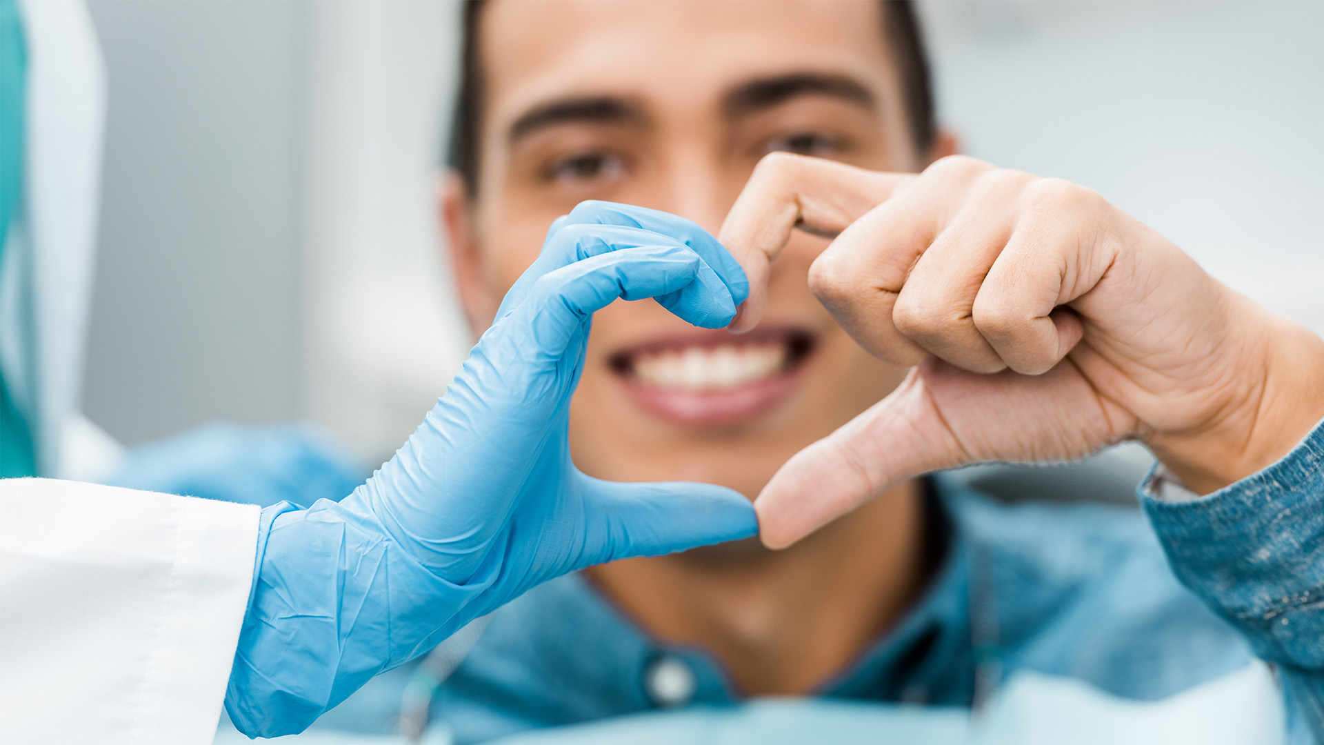 The image shows a person making a heart shape with their hands, standing behind a medical professional wearing gloves and a surgical mask, who appears to be taking a photo of them.