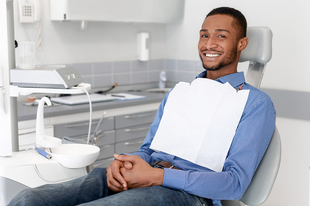 The image shows a man sitting on a dental chair with his mouth open, wearing a blue shirt, smiling at the camera, and looking directly at it.