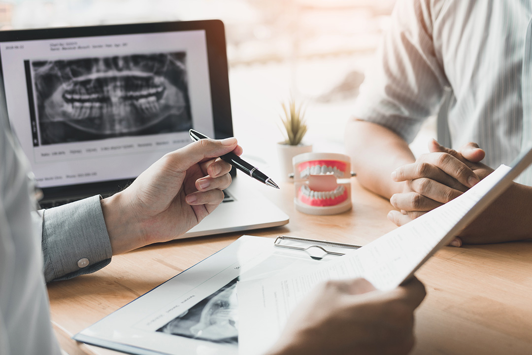 The image shows two individuals seated at a desk with a laptop, papers, and a dental X-ray displayed on a monitor. They appear to be engaged in a discussion or presentation related to dental health.