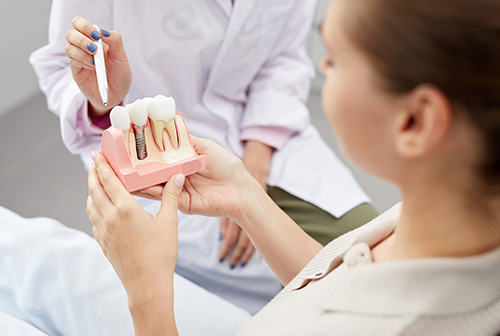 The image shows a dental professional holding an open pink mouth model with a toothbrush inside, demonstrating proper oral hygiene techniques to a patient seated nearby.