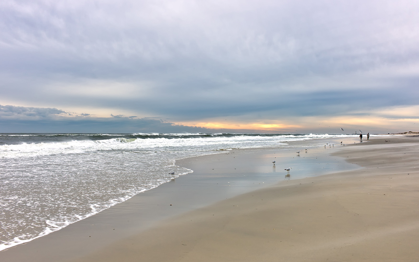 The image shows a beach scene at sunset with a clear horizon, featuring a sandy shoreline with waves gently lapping against the shore, a few birds standing on the wet sand, and a person walking along the water s edge under a cloudy sky.