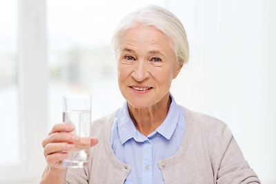 The image shows an elderly woman holding a glass of water with both hands while smiling at the camera.