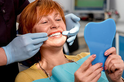 The image shows a woman sitting in a dental chair with a smile on her face, holding up a blue mouthguard, while a dentist is adjusting her teeth with a pair of tweezers.