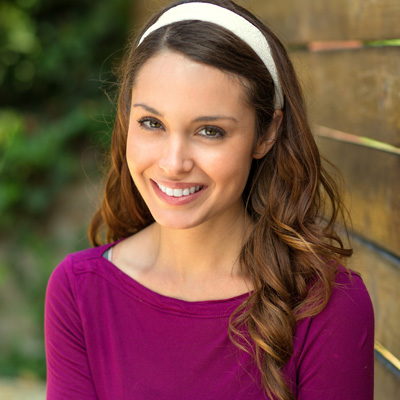 A young woman with long hair smiling at the camera, wearing a purple top and standing against a wooden fence.