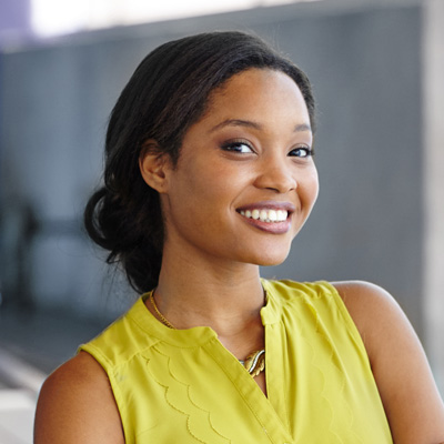 The image shows a smiling woman with dark hair, wearing a yellow top, standing against a neutral background.