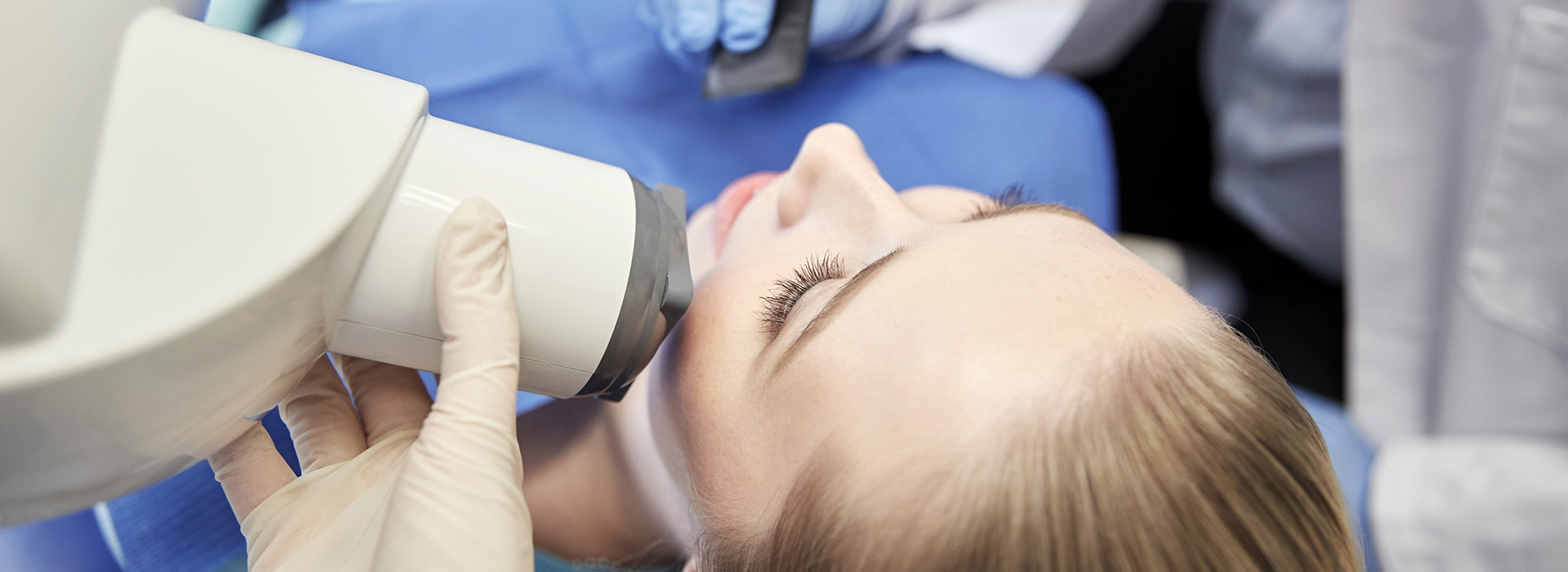 The image shows a woman receiving dental treatment under a dental microscope, with a dental professional examining her teeth closely.