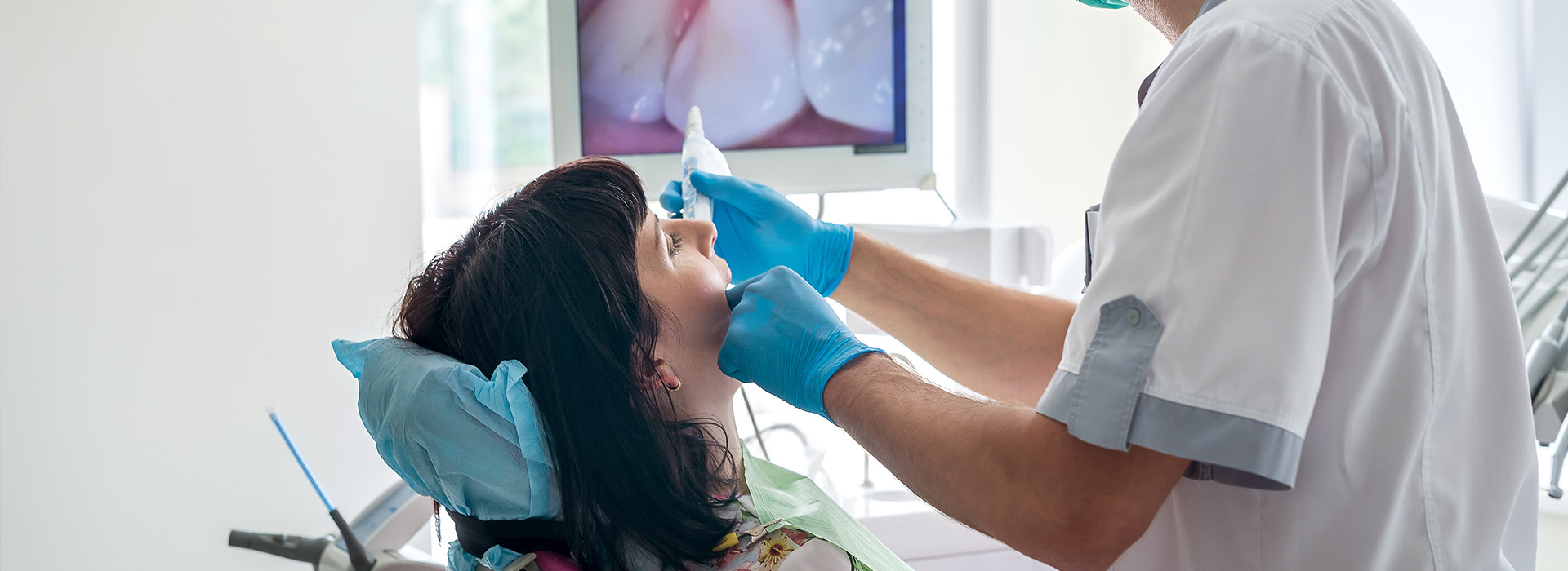 The image shows a dental professional performing a procedure on a patient s mouth, with the practitioner wearing gloves and using dental tools while seated in a clinical setting.