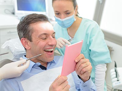 The image shows a man sitting in a dental chair with his mouth open, holding a pink card in front of him, while a female dentist stands behind him wearing a surgical mask and holding a mirror, smiling at the camera.