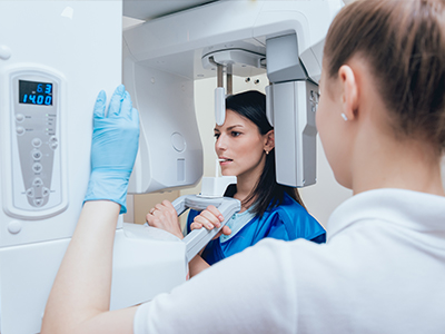 The image depicts a woman standing in front of a large medical scanner with a digital display, while another person looks on, both wearing white gloves and protective eyewear.