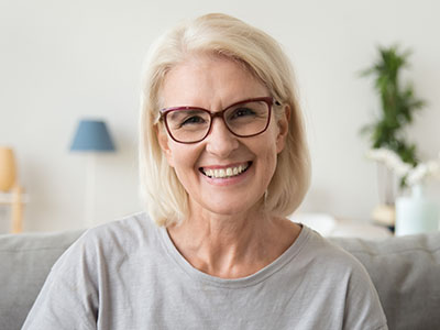 The image shows a woman with short blonde hair smiling at the camera, wearing glasses and a light-colored top, seated on a couch in an indoor setting.