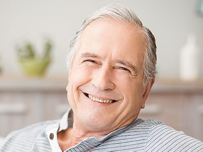An elderly man with gray hair, smiling broadly at the camera, sitting comfortably in a room with a white wall and a window.