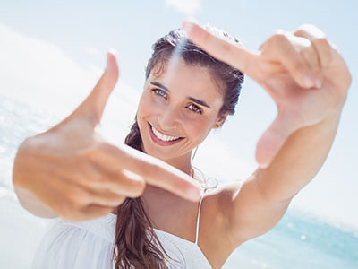 The image shows a woman taking a selfie with her fingers spread wide against a bright blue sky background.